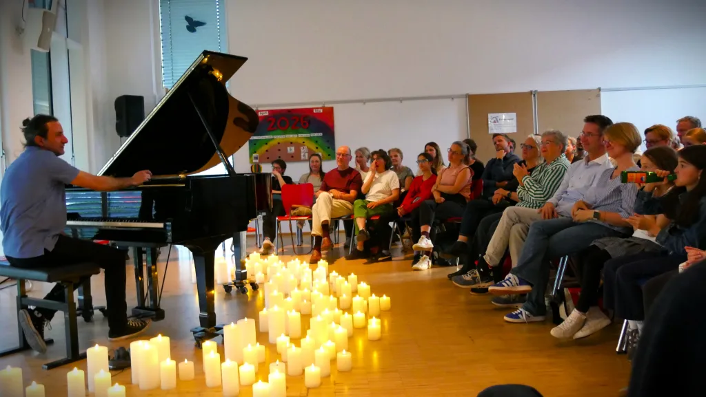 Davide Martello spielt auf einem schwarzen Flügel in der Aula der Regenbogenschule. Auf dem Boden stehen viele LED Kerzen.