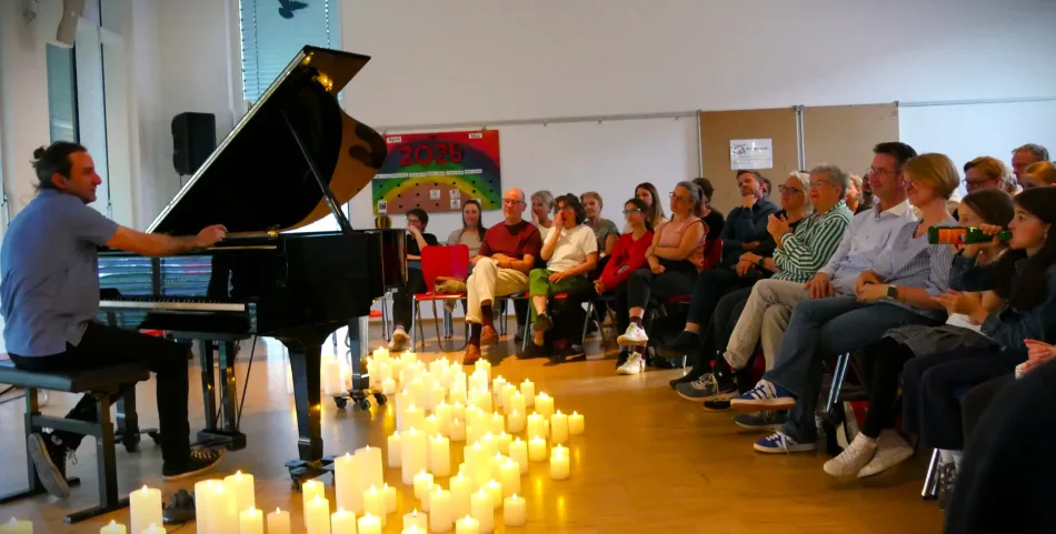 Davide Martello spielt auf einem schwarzen Flügel in der Aula der Regenbogenschule. Auf dem Boden stehen viele LED Kerzen.